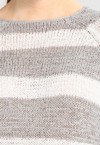Stickad tröja med rund halsringning, som har horisontella ränder i ljusbeige och grå nyanser, med ett texturerat, öppet vävsmönster.