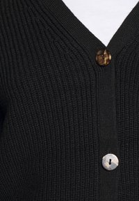 Černý žebrovaný cardigan s výstřihem do V a dvěma knoflíky: jeden s tmavým želvovinovým vzorem a druhý v světle šedé barvě s tečkovaným designem.