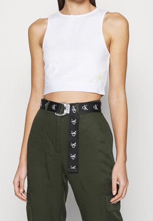Biały krótki top na ramiączkach wykonany z gładkiej tkaniny, z małym żółtym logo. W połączeniu z oliwkowozielonymi spodniami i czarnym paskiem z białym logo.