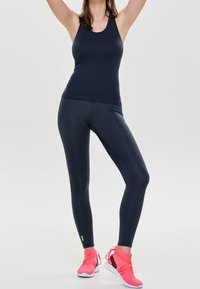 Donna che indossa un top sportivo aderente nero e leggings con scarpe da ginnastica rosa brillante, in piedi con le braccia alzate su uno sfondo neutro.