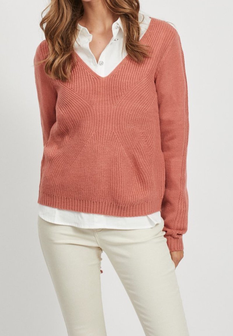 Femme portant un pull rose poussiéreux en maille à col en V superposé sur une chemise blanche à boutons avec un pantalon couleur crème.