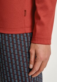 Rode katoenen langmantel bovenstuk met een zijlabel. Daaronder zijn er gemusterde onderstukken in marineblauw, rood en lichtblauwe rechthoeken die zich verticaal herhalen.