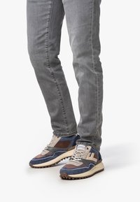 Sneaker mit einer Kombination aus marineblauen, beigen und braunen Materialien, mit einer strukturierten Gummisohle und Netzpanelen für Atmungsaktivität.