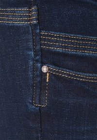 Mörkblå denimjeans med kontrasterande gult sömnad och en liten metallnitar nära hörnet av framfickan.