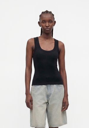 Femme aux cheveux tressés portant un débardeur noir sans manches et un short en jean clair longueur genou sur un fond clair uni.