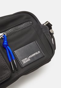 Černá nylonová crossbody taška s zipovou kapsou, modrým šňůrovým akcentem a embosovanou koženou etiketou s brandingem "KARL LAGERFELD JEANS".