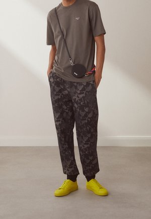 Person trägt ein olivgrünes T-Shirt, dunkle Camouflage-Jogginghosen, leuchtend gelbe Turnschuhe und eine schwarze runde Umhängetasche mit buntem Riemen.