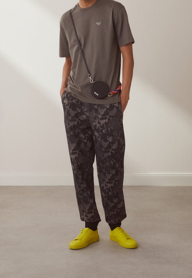 Personne portant un t-shirt vert olive, un jogging camouflage foncé, des baskets jaunes vives et un sac bandoulière rond noir avec une sangle colorée.