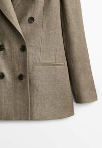 Veste blazer kaki à double boutonnage avec des boutons noirs, des revers crantés et une poche latérale unique ; fabriquée en tissu texturé.