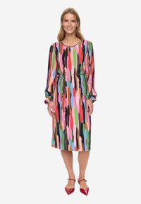 Kleurrijke jurk met patroon, lange mouwen, een losse pasvorm, ceintuur in de taille en een zoom tot op de knie. Rode schoenen maken de outfit compleet.