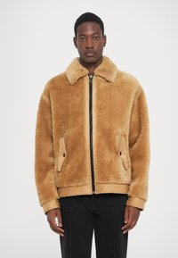 Tan faux shearling jakke med lynlås foran, spidse kraver og sidelommer. Har en blød tekstur og et afslappet fit design.