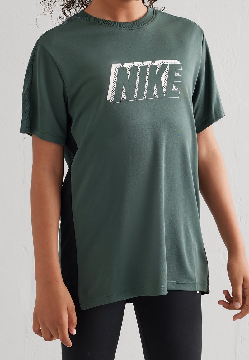 Persona vestida con una camiseta deportiva Nike de color verde oscuro y leggings negros, de pie frente a una pared clara y lisa.