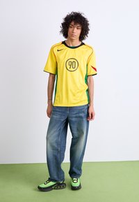 Yellow sports jersey with a round logo and number 90, featuring green and red accents; paired with loose blue jeans and green sneakers.