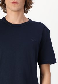 Camiseta de algodón azul marino de manga corta, con cuello redondo y un sutil detalle del logo "1962" en el lado izquierdo del pecho. Textura suave.