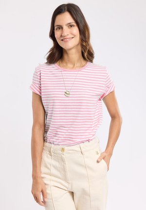 Femme aux cheveux bruns mi-longs, portant un t-shirt rayé rose et blanc, un pantalon beige et un collier avec pendentif coquillage, souriante.