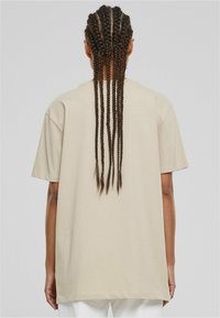 Camiseta oversized beige de manga corta, hecha de algodón suave. Corte relajado con cuello redondo, mostrando cabello trenzado en la parte de atrás.