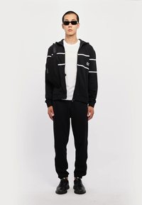 Svart zip-up hettegenser med hvite striper, hvit T-skjorte, svarte joggebukser og solbriller. Modell som holder en liten svart veske. Sneakers på føttene.