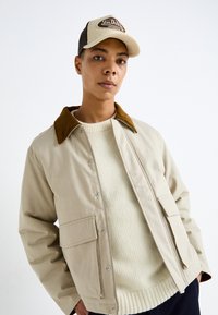 Chaqueta de tela beige con un cuello de pana marrón, botones a presión y bolsillos frontales, llevada sobre un suéter de punto crema. Gorra ajustable en la cabeza.