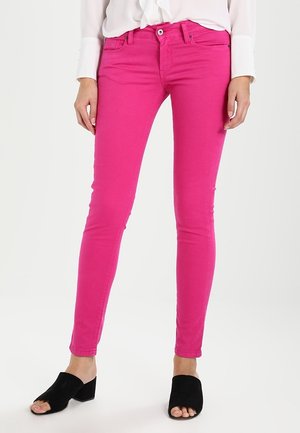 Woman wearing bright pink skinny jeans, white blouse, and black open-toe low-heel shoes, standing against a plain light background.