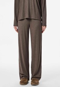 Osoba u slobodnim, smeškastim pletenim hlačama i dugim rukavima u istom tonu, uparenim s smeškastim zamščenim papučama na bijeloj pozadini.