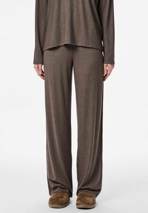 Osoba u slobodnim, smeškastim pletenim hlačama i dugim rukavima u istom tonu, uparenim s smeškastim zamščenim papučama na bijeloj pozadini.