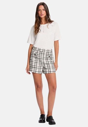 Femme portant un haut blanc à manches courtes avec des détails à boutons et un short à carreaux noir et blanc, assorti à des mocassins noirs.