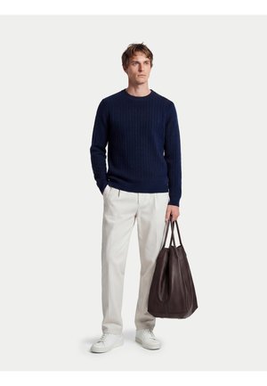 Mann trägt einen dunkelblauen Pullover, weiße Hose, weiße Sneakers, hält eine große dunkelbraune Ledertasche und steht vor einem einfarbigen Hintergrund.