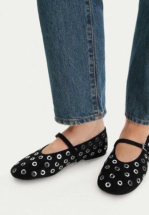 Füße in schwarzen Mary Jane Ballerinas mit silbernen Ösen, kombiniert mit gekürzten blauen Jeans.