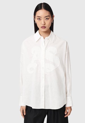 Jeune femme aux cheveux noirs raides portant une chemise blanche à boutons surdimensionnée avec un motif "A3" en relief et un pantalon noir, sur fond gris clair.
