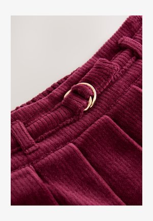 Tessuto in velluto a coste color bordeaux con righe verticali, caratterizzato da una chiusura in metallo colore oro e un dettaglio con passante per cintura. Vista ravvicinata.