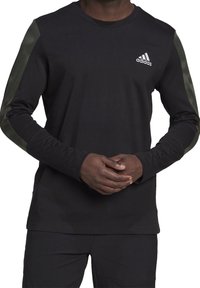 Muže v černém tričku s dlouhými rukávy značky Adidas s tmavě zelenými pruhy na rukávech, s rukama skloubenýma vpředu, v odpovídajících černých kalhotách.