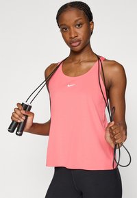 Ροζ αθλητικό τοπ χωρίς μανίκια με στρογγυλή λαιμόκοψη, διακοσμημένο με μικρό λευκό λογότυπο Nike. Το μοντέλο κρατάει ένα μαύρο σχοινάκι άλματος με υφασμάτινες λαβές.