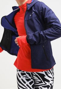 Nike takki tummansinisellä värillä ja tekstuurilla, yhdistetty punaisen pitkähihainen paidan ja mustavalkoisten kuviollisten trikoojen kanssa, joissa on vetoketju.