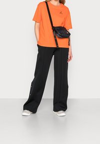 Pomarańczowy bawełniany t-shirt z logo, w połączeniu z czarnymi szerokimi spodniami z guzikami zatrzaskowymi. Czarna torba na ramię z paskiem.