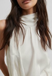 Beżowa bezrękawnikowa bluzka z wysokim kołnierzem, wykonana z miękkiego materiału i z falbaniastymi detalami z przodu, co sprawia, że ma luźny krój.