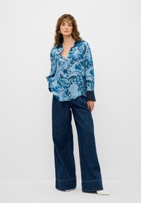 Camicia blu con motivo paisley, colletto e maniche lunghe, abbinata a jeans in denim scuro a gamba larga e scarpe bianche a punta.