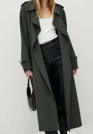 Woman wearing long dark green coat over white top and black pants, holding a black woven handbag by her side.