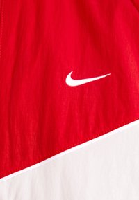 Rød vindjakke med en glatt tekstur, med en hvit Nike-logo og kontrasterende hvit underdel. Inkluderer pene sømdetaljer.