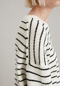 Gestreifter Pullover mit lockerer Passform, der schwarze Streifen auf einem weißen Strickhintergrund zeigt. Strukturiertes Material mit sichtbaren Nähdetails.