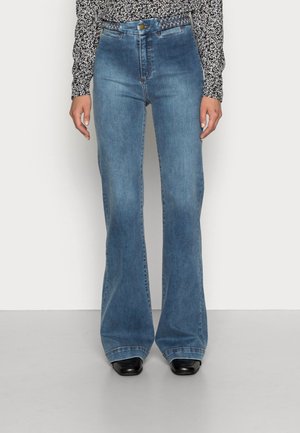 Kobieta w niebieskich dzwonowatych jeansach z wysokim stanem, z plecionym paskiem, czarnych butach i czarno-białej koszuli z długim rękawem w wzór.