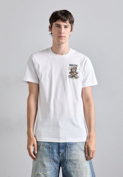 Witte katoenen T-shirt met een afbeelding van een beer en tekst op de linkerborstzijde, gecombineerd met lichtblauwe acid-washed jeans.