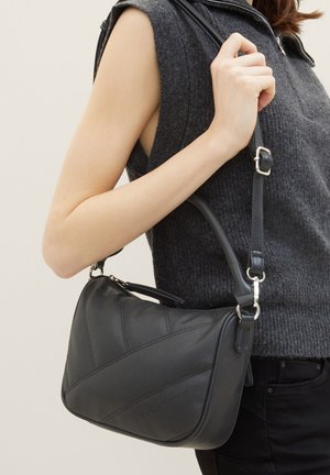 Sac à main noir texturé avec des coutures diagonales, une seule bandoulière réglable et des détails en métal argenté, tenu par une personne portant un haut gris.