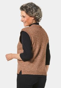 Bruine gebreide vest met mouwloos ontwerp, ribbelrand, zijsplitten en textuurstof, gedragen over een zwart shirt met lange mouwen.