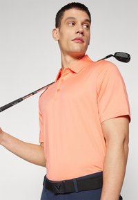 Kortermet polo-skjorte i lysoransje, med krage, tre knapper og teksturert stoff. Båret med svart belte og golf kølle.