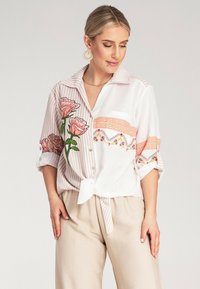 Biała koszula z guzikami, w różowy paski z haftem kwiatowym. Posiada zawiązywany przód, podwinięte rękawy i akcenty koronkowe.