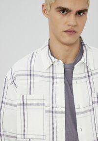 Chemise à carreaux blanche avec des accents violets, tissu texturé, design à boutons, dotée d'une poche sur la poitrine et portée sur une chemise grise.