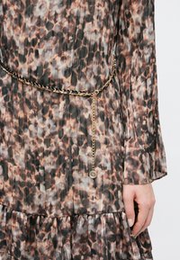 Πλισέ φόρεμα με λεοπάρ μοτίβο, χρυσή ζώνη αλυσίδα με υφαντή μαύρη κορδέλα και κρεμαστό μενταγιόν, το χέρι ακουμπάει στο πλευρό.