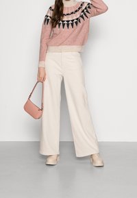 Rosa Strickpullover mit schwarz-weißer geometrischer Musterung, kombiniert mit weit geschnittenen cremefarbenen Hosen und hellbeigen Plateauschuhen, während eine rosa Tasche gehalten wird.