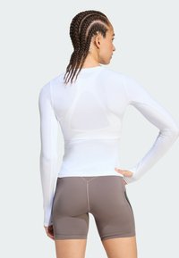 Λευκή αθλητική μπλούζα με μακριά μανίκια, εφαρμοστή σχεδίαση και υφασμάτινες λεπτομέρειες, συνδυασμένη με καφέ εφαρμοστά σορτς που έχουν αντίθετη ραφή.