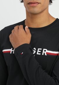 Mężczyzna w czarnej bluzie z długim rękawem z poziomymi paskami w kolorach czerwonym, białym i niebieskim oraz częściowym napisem "HILFIGER" na klatce piersiowej, opiera rękę na ramieniu.